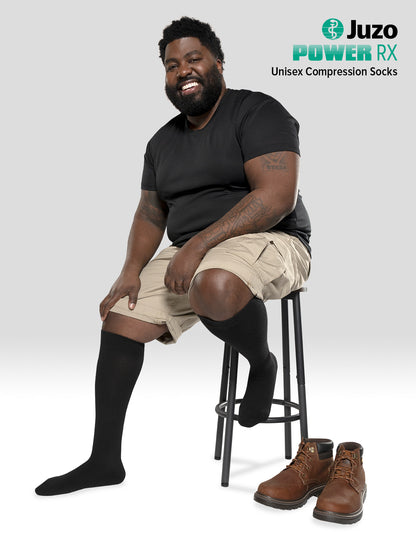 Man wearing Juzo Power RX compression socks while sitting on a stool, showing the branded logo – Compressionsale compression wear for leg support.