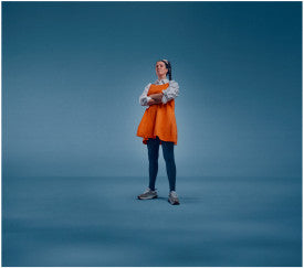 Person wearing an orange dress and blue jeans standing against a solid blue background